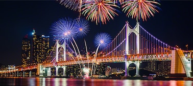 Fireworks in a night sky over an illuminated bridge reflected in a river below