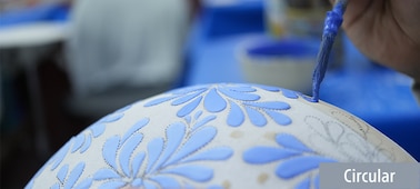 A sample image showing a craftsman adding patterns to a white jar using a brush and blue paint. The focus is on the brush, while the foreground of the jar and the background blurred