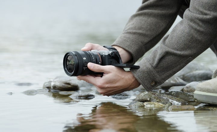 A usage image showing a user holding the α7CII with the FE 24-50mm F2.8 G near the surface of a river. The user is shooting from a low position